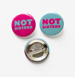 Two pinback buttons Button on the left reads NOT SISTERS with a teal background and magenta pink text. Right sided button reads NOT SISTERS in lteal with a magenta pink. Third button shows metal pinback.