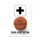 Round warm brown colored 1.25" pinback that reads NUT ALLERGY in a dark  brown text. On a white backing card, black plus sign at the top. Below reads: Social Alert Button, with tiny text beneath it which says, for common physical & psychological situations