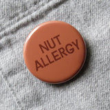 Round warm brown colored 1.25" pinback that reads NUT ALLERGY in a dark  brown text. Button is pinned to a gray flannel shirt.