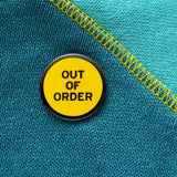 Round enamel pin that says OUT OF ORDER. Gunmetal text and outline on a yellow background.