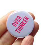  Round light gray button that reads OVER THINKINER in two lines in pink text. Badge is held in a hand.