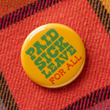 a round marigold yellow-orange pinback button that reads PAID SICK LEAVE in green text, and FOR ALL in red text. Button is pinned to a flannel shirt