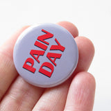 Round gray pinback button that reads PAIN DAY in a chunky red font with black drop shadow. Button is head in a hand by the fingers.