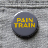 Round gray pinback button that reads PAIN TRAIN in yellow text. Button pined to gray flannel pocket.
