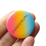 Round 1.25" pinback button with a pink yellow blue diagonal ombre that reads PANSEXUAL in a thin black san serif font.  Badge is held in hand.