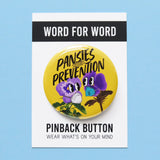 Round butter yellow 2.25" pinback button that reads Pansies for Prevention in a black text over the image of two purple pansies wearing, one wearing a KN95 mask, the other a Flo Mask Pro with cute cartoon eyes, and photo stems. Button is on a Word For Word branded backing card that reads Wear What Is On Your Mind