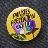 Round butter yellow 2.25" pinback button that reads Pansies for Prevention in a black text over the image of two purple pansies wearing, one wearing a KN95 mask, the other a Flo Mask Pro with cute cartoon eyes, and photo stems. Pinned to black denim.