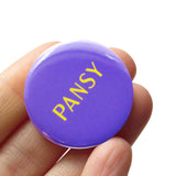 A purple pinback button that reads PANSY in a vintage yellow font. Badge is held in a hand.