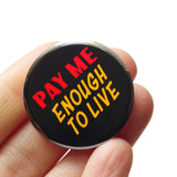 A round black 1.25" pinback button that reads PAY ME ENOUGH TO LIVE in red and yellow text. Badge is held in a hand.