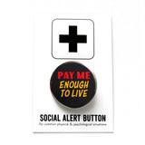 A round black 1.25" pinback button that reads PAY ME ENOUGH TO LIVE in red and yellow text. Badge is pinned to a Social Alert Button backing card.