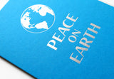 A bright blue A2 greeting card with a silver letterpress imprint of a planet earth illustration that reads PEACE ON EARTH, close up of the card at an angle.