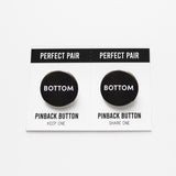 Two pinback buttons on a backing card with a center perforation to tear & share with a friend. Button the left reads BOTTOM. Button on right reads BOTTOM. Both buttons are black with white text.
