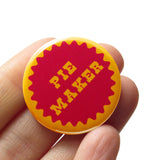 A round golden brown pie crust color 1.25" pinback button with a jammy berry center that reads PIE MAKER in golden cut out letter. Button is held in a hand.