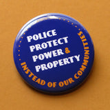 A round navy blue pinback button that reads Police Protect Power & Property, Instead of Our Communities in white & golden yellow text. Button is on a warm sand oat colored background.