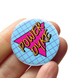Round light pinback button with a lavender grid pattern, and magenta pink triangle reads POWER DYKE in golden yellow text. Badge is held in a hand.