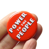 Round red-orange pinback button with white art deco text that reads POWER TO THE PEOPLE with a black drop shadow. Button is held in a hand.