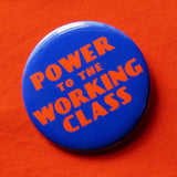 A round bold blue pinback button that reads POWER TO THE WORKING CLASS in red-orange text,  an art deco font. Button on a red-orange fabric.