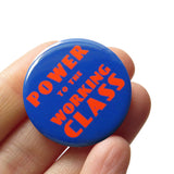 Round blue pinback button with red-orange text that reads POWER TO THE WORKING CLASS in an art deco font. Button is held in a hand.
