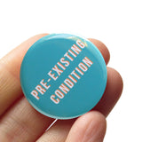Round pinback button that says PRE-EXISTING CONDITION. White text on a blue background. Button is held in a hand.