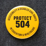 Round yellow pinback button that reads PROTECT 504 in the center, and Accessibility & Disabled Civil Rights along the top, and In Education & Healthcare along the bottom. Pinned to black denim.