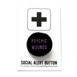 Round black pinback button that reads PSYCHIC WOUNDS in a thin lavender font. Pinned to a Social Alert Button backing card.