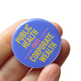 A round pinback button with a scrubs blue-purple background that reads PUBLIC HEALTH OVER CORPORATE WEALTH in yellow and neon red. Button is on a Social Alert Button backing card.