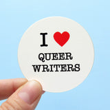 A round white sticker that reads I LOVE QUEER WRITERS with love indicated by a red heart. Send up by a hand, in the lower left corner, against a light blue background.