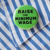 Round green pinback button that reads RAISE THE MINIMUM WAGE in black text. Button is pinned to a blue & white stripped shirt like a fast food worker might be made to wear at work.