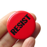  round bright red pinback button that reads RESIST in black type. Button is held in a hand.