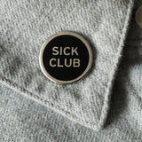 Round enamel pin that says SICK CLUB. Silver text and outline on a black background.