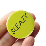 round lime green pinback button that reads SLEAZY in a thin black font. Button held in a hand, by fingers.
