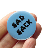 Round light blue 1.25" pinback button that reads SAD SACKin a chunky black font. Button is held in  a hand.