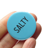 Sea salty blue, round pinback button that reads SALTY in black, thin tall san serif font. Button is held in a hand.