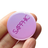 ound light lavender to pink ombre 1.25" pinback button with that reads SAPPHIC in a dark pink san serif font.  Button is held in a hand.