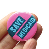 A round pink pinback button with light aqua text and dark teal boxes that reads SAVE MEDICAID. The buttons held in a hand.