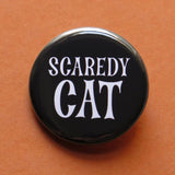 A round black pinback button that reads SCAREDY CATin a spooky white font. Button is on a rust colored background