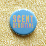 Round, visually textured light blue pinback button that reads SCENT SENSITIVE in a deep cream color with pink drop shadow. Button is on a butter yellow microfiber cloth