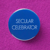 Bold bright blue round pinback button that reads SECULAR CELEBRATOR in thin white sans serif text.  Button is pinned to a raspberry sweatshirt.