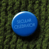 Bold bright blue round pinback button that reads SECULAR CELEBRATOR in thin white sans serif text.  Button is pinned to a green sweater