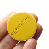 A round yellow 1.25" pinback button that rads SENSITIVE in thin black text. Badge is held in a hand.