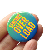 A round green to blue ombre 1.25" pinback button that reads SENSORY OVERLOAD in bright yellow text with a pink drop shadow. Button is held in a hand.