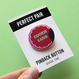 A button that looks like a chocolate cookie  with a bite missing & reads COOKIE EATER in mint green. Button is on a backing card that reads PERFECT PAIR, Pinback Button, Share one, and is held in the corner by a thumb & forefinger.