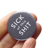 A round, dark gray 1.25" pinback button that reads SICK OF THIS SHIT in white text. Button is held in a hand.