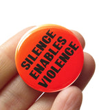 A round pinback button with an orange-red background, that reads SILENCE ENABLES VIOLENCE in black text. Button is held in a hand.