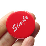 Round red pinback button that reads SINGLE in a white script font. Badge is held in a hand.