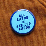 Round light blue pinback button with a dark blue outline & dark blue text that reads ALL LABOR IS SKILLED LABOR. The button is on a rich warm caramel colored sweatshirt.
