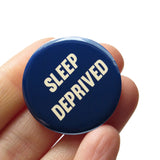 Round navy blue pinback button that reads SLEEP DEPRIVED in cream slanted text. Badge is held in a hand.