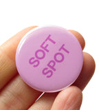 Round light pink 1.25" pinback button that reads SOFT SPOT in dark pink. Badge is held in a hand.