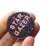 Round pinback button that reads, STAR GAZER. Salmon pink text with a light purple drop shadow, set on a background of outer space, full of stars. The button is held in a hand.
