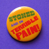 A round yellow-orange pinback button that reads STONED AND IN TERRIBLE PAIN! in green. purple & red text.  Button is on a purple background.